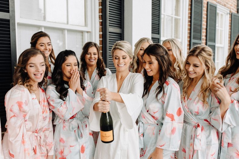 bride opening bottle of champaign with bridesmaids in floral robes surrounding her