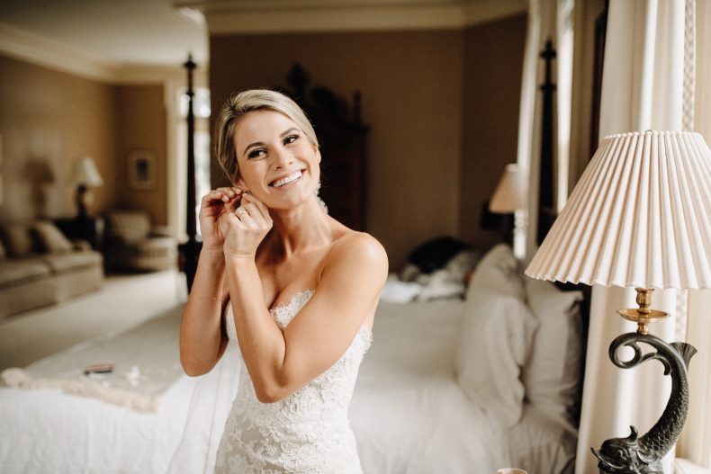 blonde bride in lace fitted wedding gown putting in earrings